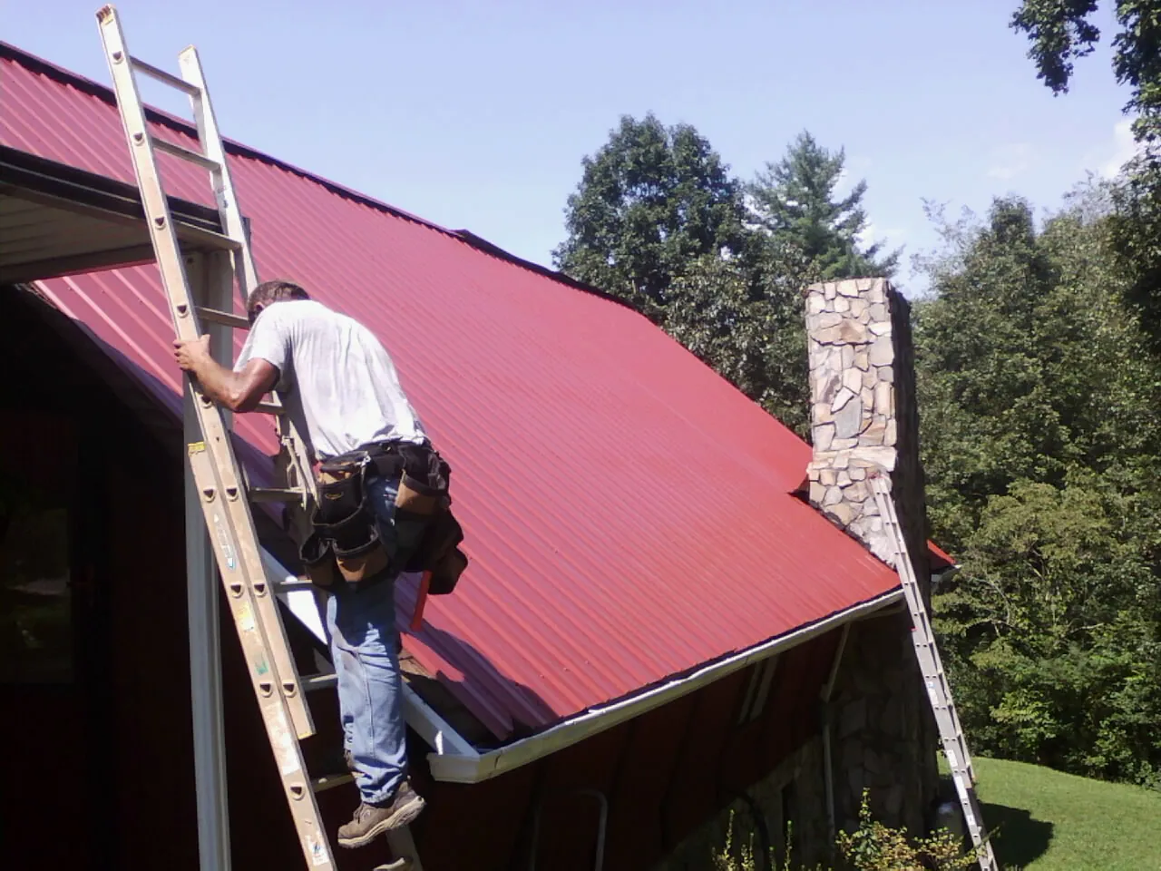 Roofer on a ladder beside a red metal roof and stone chimney — Soffit & Fascia Repair by Roofing Brothers in Black Mountain